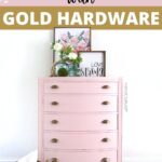 A pink vintage dresser with gold hardware, floral art, and decor on top, against a white wall.