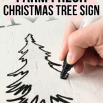 Hand drawing a Christmas tree outline with black marker on a white wooden surface; text reads DIY Farm Fresh Christmas Tree Sign.