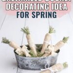 A metal bucket labeled Flowers & Garden holds twine-wrapped faux carrots with greenery tops.