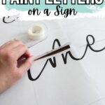 Hand cutting stencil outline of cursive letters on white paper with tape dispenser nearby.