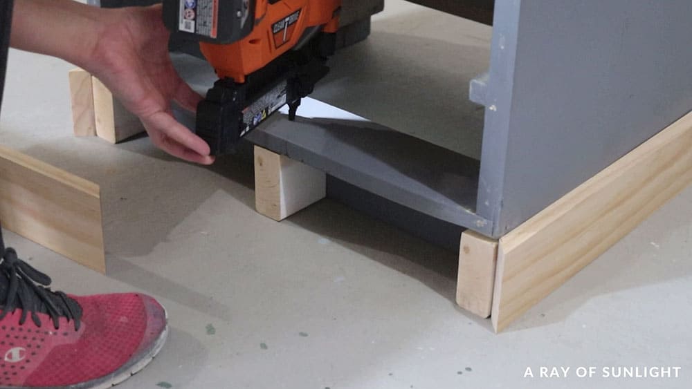 Using a brad nailer to attach supports and wood trim to the bottom of the cabinet.