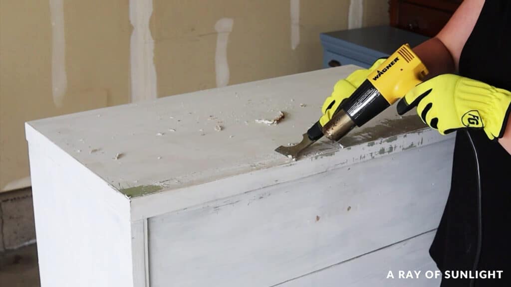 a person pointing a heat gun to the wood surface while scraping the paint with a putty knife