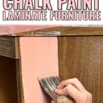 A hand painting pink chalk paint on a brown laminate furniture piece with a brush.