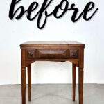 A small vintage wooden sewing table stands in front of a white wall with before written in large script.