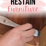 Hand using a brush to apply wood stain to furniture, with text How to Restain Furniture above.