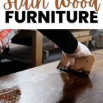 A gloved hand spreads wood stain on a wooden surface with a flat tool.
