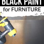 A spray gun applies black paint to a wooden surface