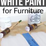 Jars of white paint and brushes on a wooden surface with paint samples labeled for furniture use.