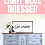 A light blue painted dresser with a wooden top, flowers, and a framed “I love you more” sign above it.