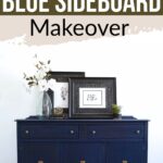 A dark glazed navy blue painted sideboard with framed art and a vase of white flowers on top, against a white wall.