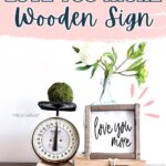 A wooden sign with love you more, books, a vintage scale, and a vase with flowers on a wooden shelf.
