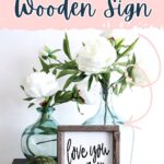 Framed “love you more” sign with white flowers in glass vases on a table, books, and green moss ball.