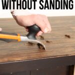 A person uses a scraper to remove paint from a wooden surface without sanding.