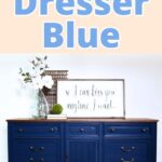 A dark blue dresser with brass handles, a vase, and a framed sign on top against a white wall.