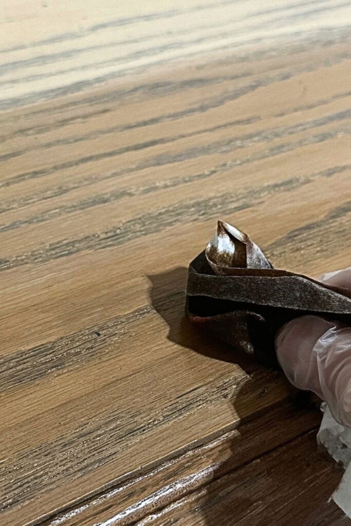 A gloved hand applies wood stain to a wooden surface using a lint free rag