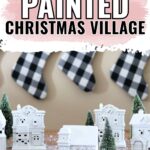 White miniature Christmas village houses with small trees in front; plaid stockings hang in the background.