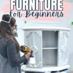 Woman in mask uses a paint sprayer on a white cabinet; text reads Flipping Furniture For Beginners.