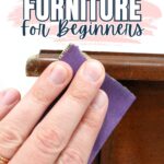 Hand sanding the edge of wooden furniture with purple sandpaper; text reads Sanding Furniture For Beginners.