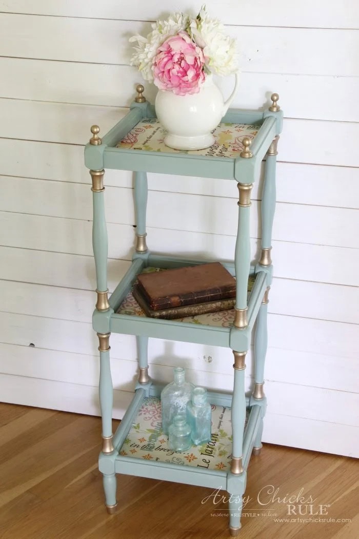 Light blue three-tier shelf with fabric decoupage holds a vase, books, and glass bottles