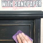 Hand using sandpaper to distress the dark-painted edge of a wooden furniture piece. Text reads: How to Distress Furniture With Sandpaper.