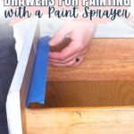 Person applying blue painter’s tape to the edge of a wooden drawer before painting.