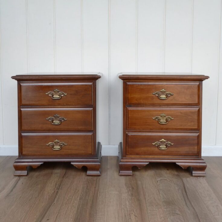 Two wooden bedside tables with three drawers each and brass handles, placed on a wooden floor against a white wall.
