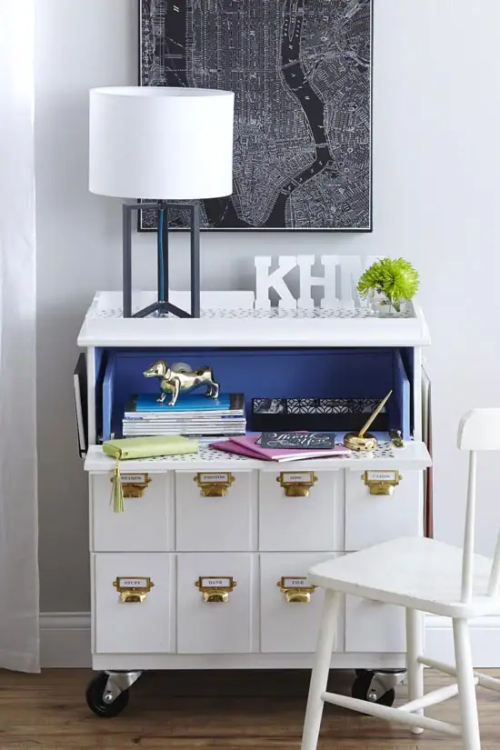 White rolling cabinet with drawers, books, lamp, and decor against a gray wall with a map art above.