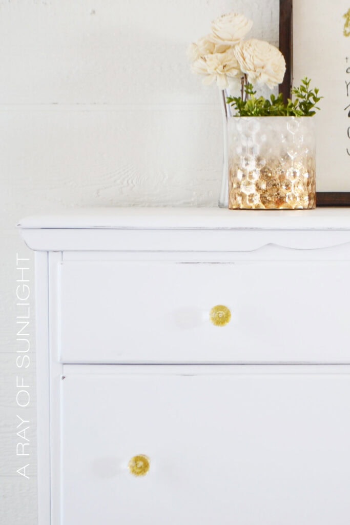 Close-up of white dresser with gold and clear knobs, topped with a gold vase and flowers
