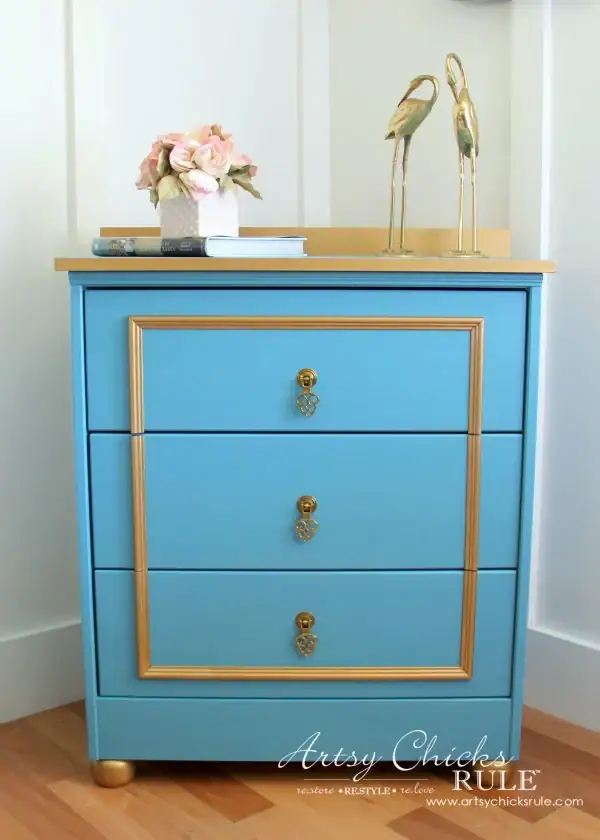 Blue dresser with gold accents, topped with a pink flower arrangement, books, and two bird figurines.