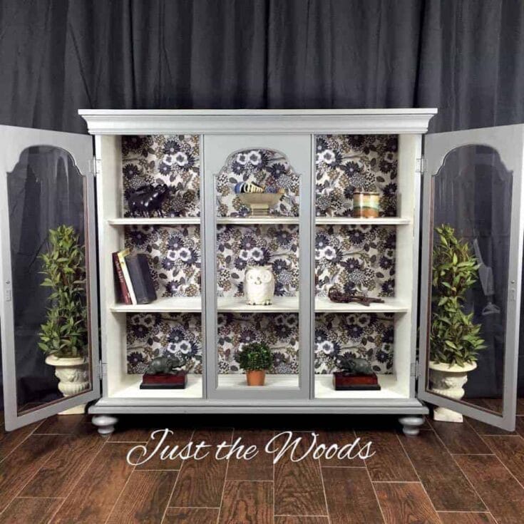 Gray cabinet with glass doors, floral wallpaper, books, plants, and decor.