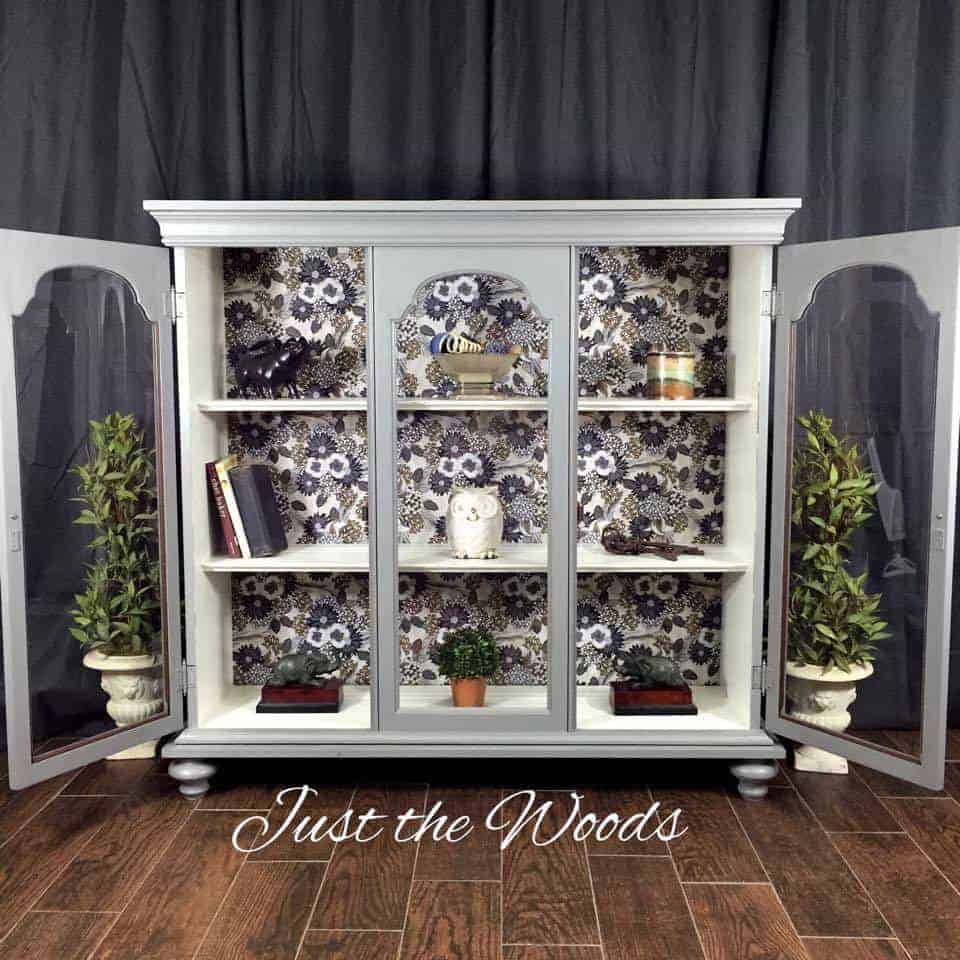 Gray cabinet with glass doors, floral wallpaper, books, plants, and decor.