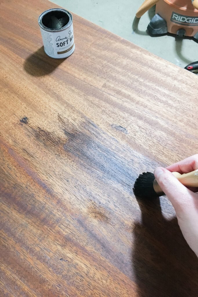 Hand applying dark wax to a wooden surface with a brush; wax container sits nearby on the table.