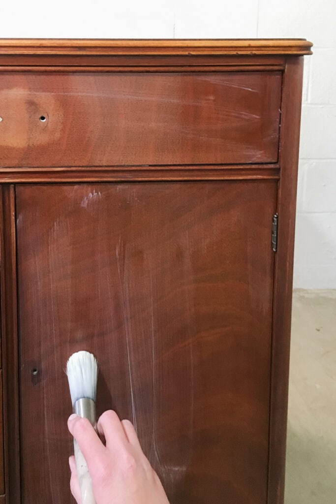 A hand uses a brush to apply a clear primer to a wooden cabinet door.