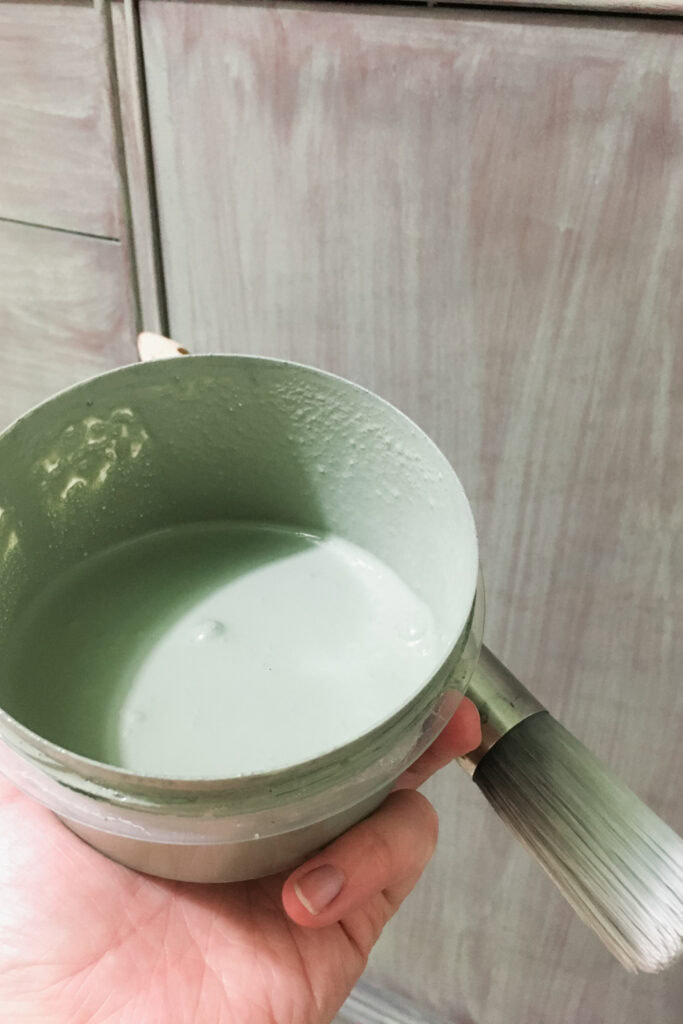 A hand holds a paint cup filled with pale green paint and a paintbrush in front of a painted cabinet.