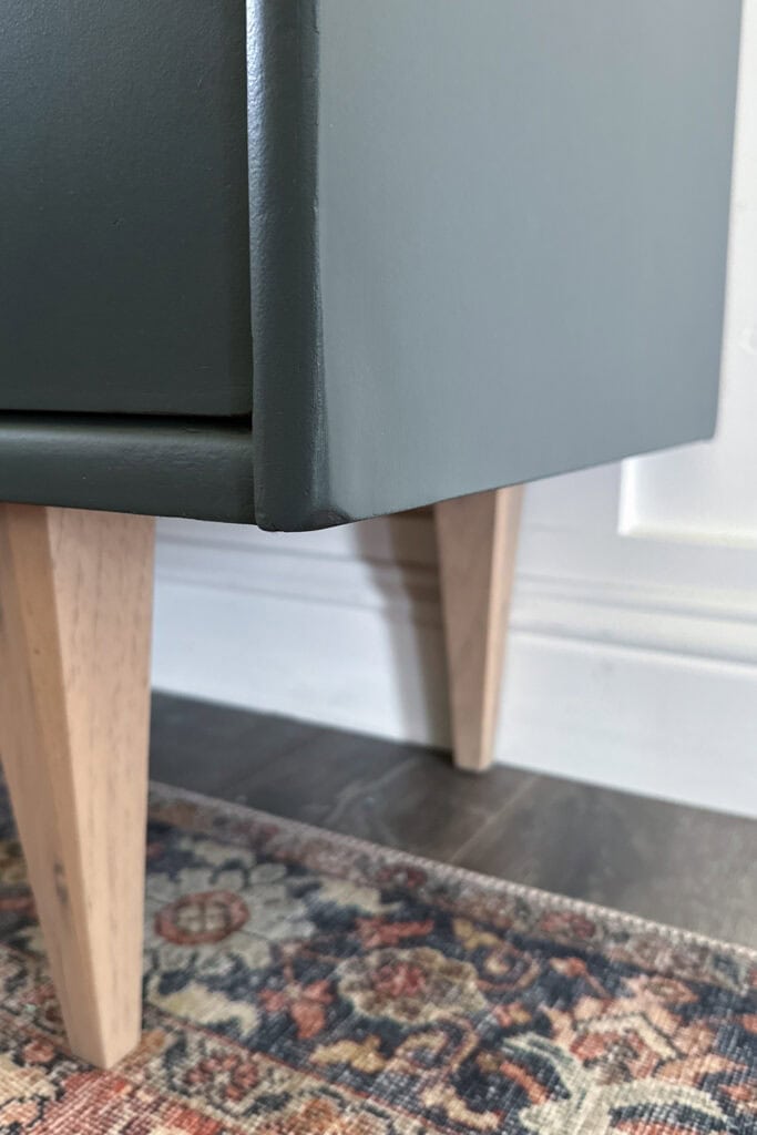 Close-up of a cabinet with light wooden legs on a patterned rug and dark floor by a white wall.
