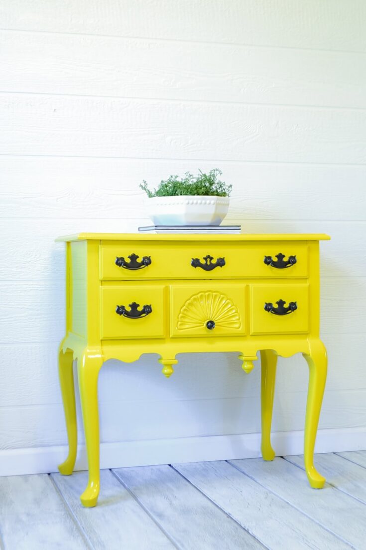Bright yellow vintage side table with ornate details and a potted plant on top, against a white wall.