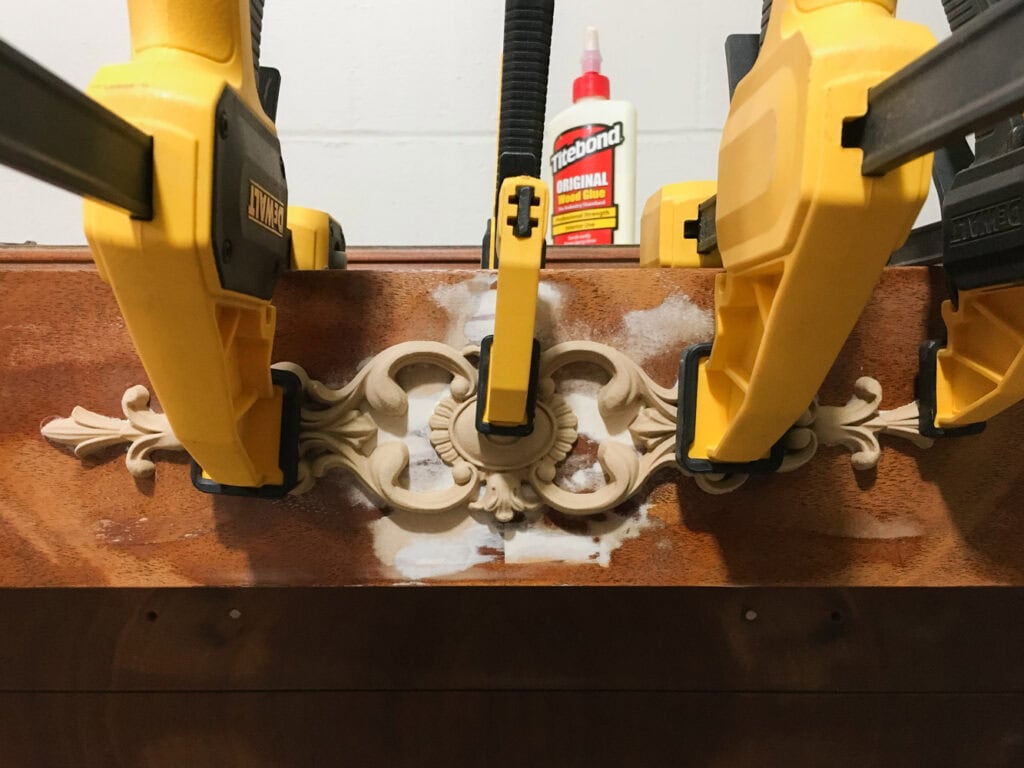 Yellow clamps hold a decorative wood applique on furniture, with wood glue and filler visible nearby.