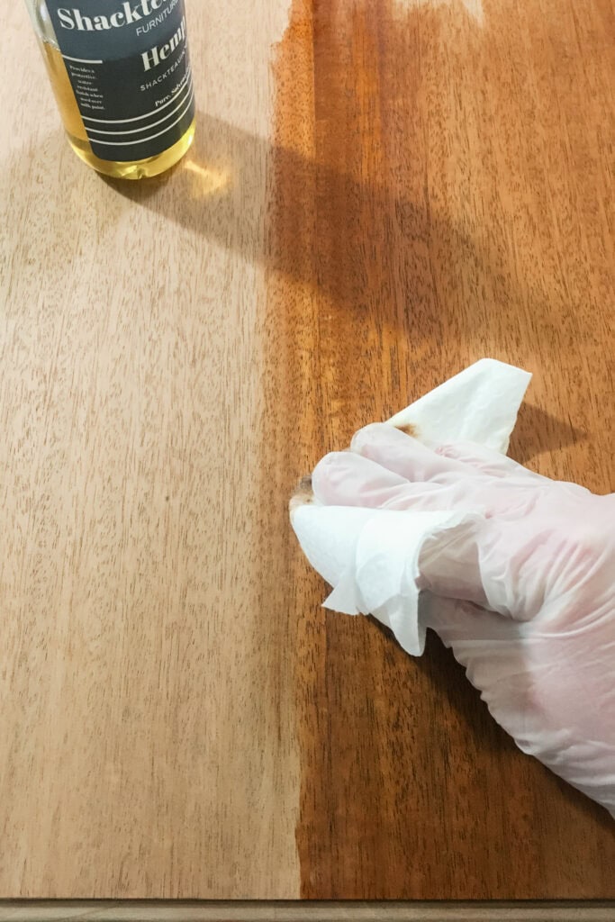 A gloved hand wipes oil on wood, showing a contrast between treated and untreated sections.