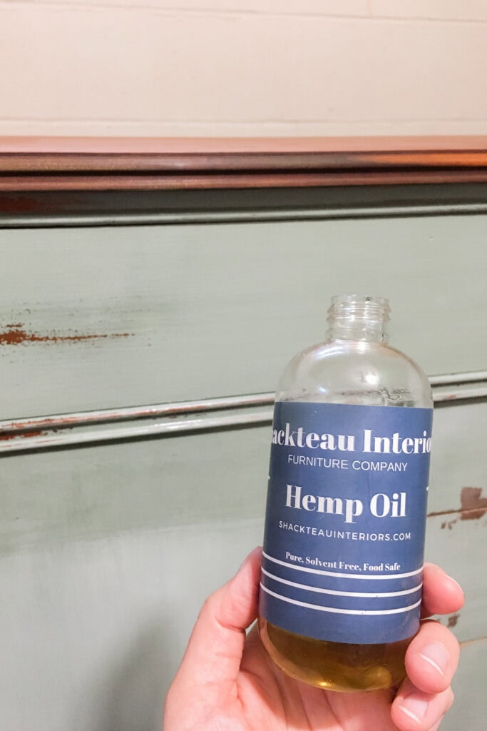 Hand holding a bottle of Shackteau Interiors hemp oil in front of a distressed green piece of furniture