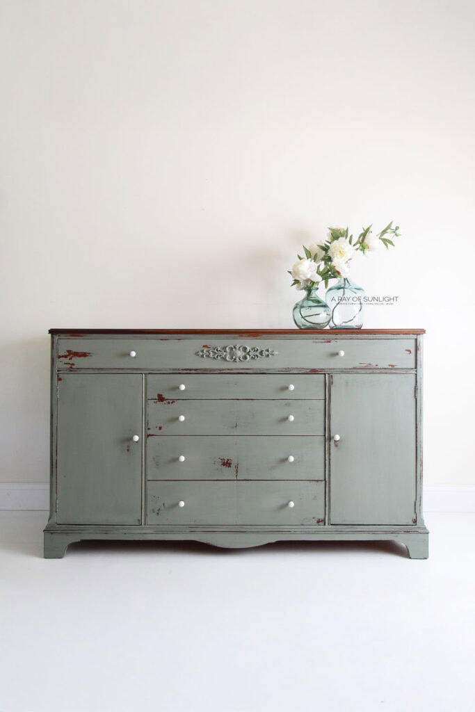 A vintage green buffet with white knobs and a vase of white flowers on top against a plain wall.