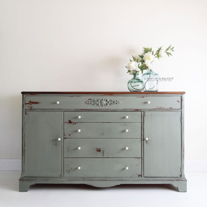A vintage green buffet with white knobs and a vase of white flowers on top against a plain wall.