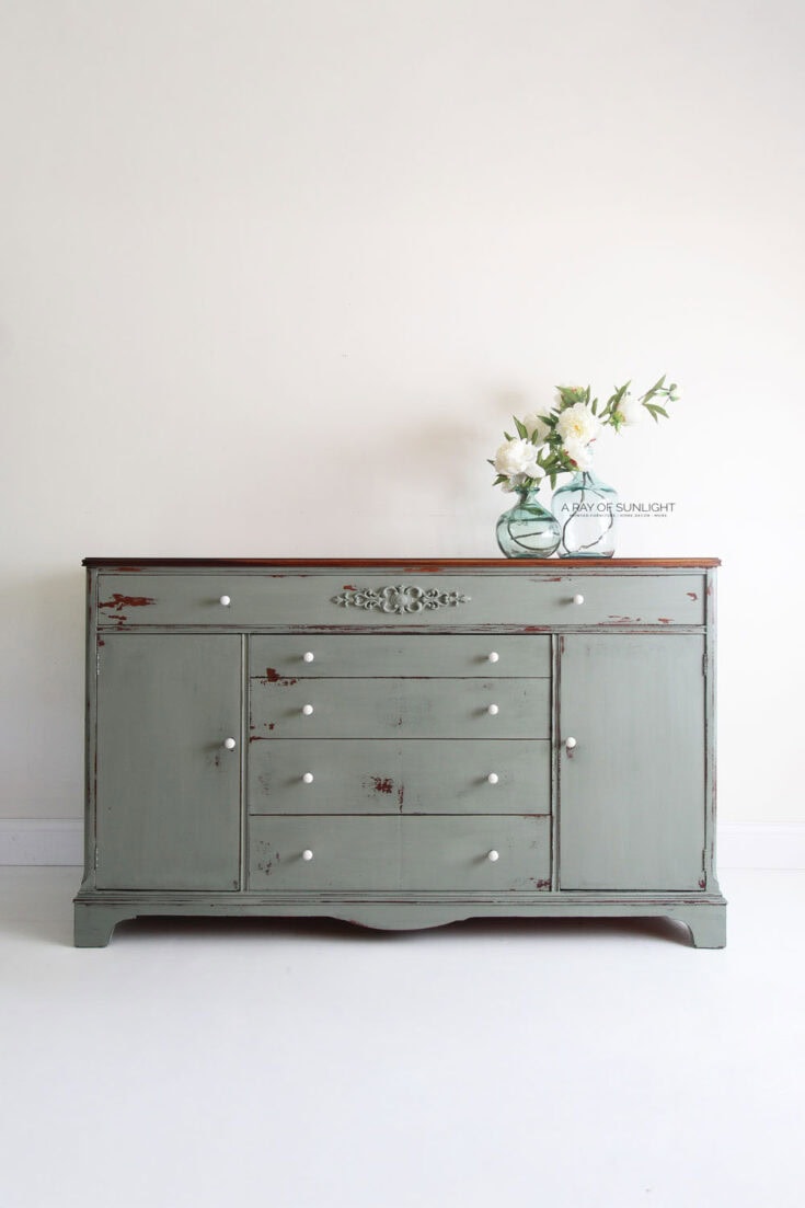 A vintage green buffet with white knobs and a vase of white flowers on top against a plain wall.