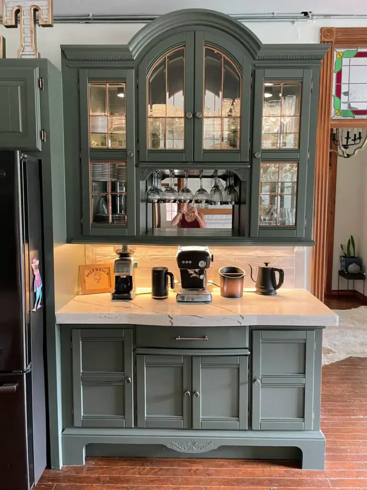 Dark green kitchen hutch with glass cabinets, coffee maker, appliances, and hanging glasses.