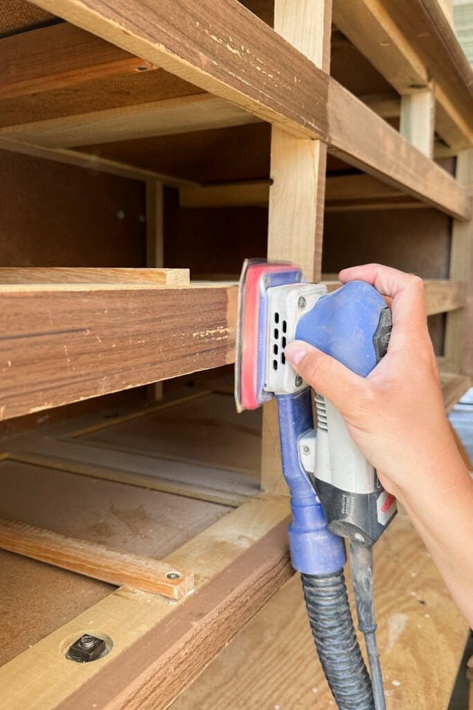 A person uses an electric sander to smooth wooden shelves inside a partially built cabinet.