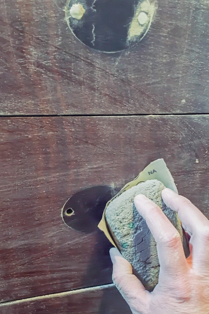 A hand is sanding a dark wooden surface with filled drawer holes
