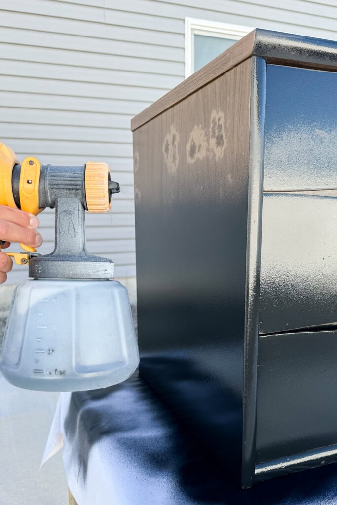 A hand holds a paint sprayer, painting a black dresser outdoors near a gray building.