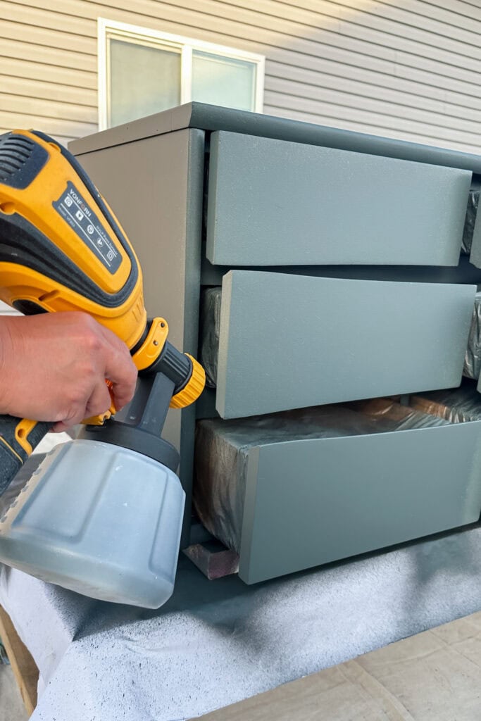 A person uses a paint sprayer to paint green drawers on a dresser outdoors.