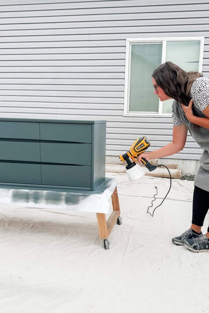 Woman spray painting a teal dresser outdoors, beside a gray house with a window.