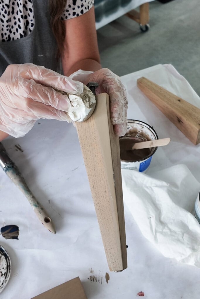 Person wearing gloves staining a wooden furniture leg with a cloth at a work table.