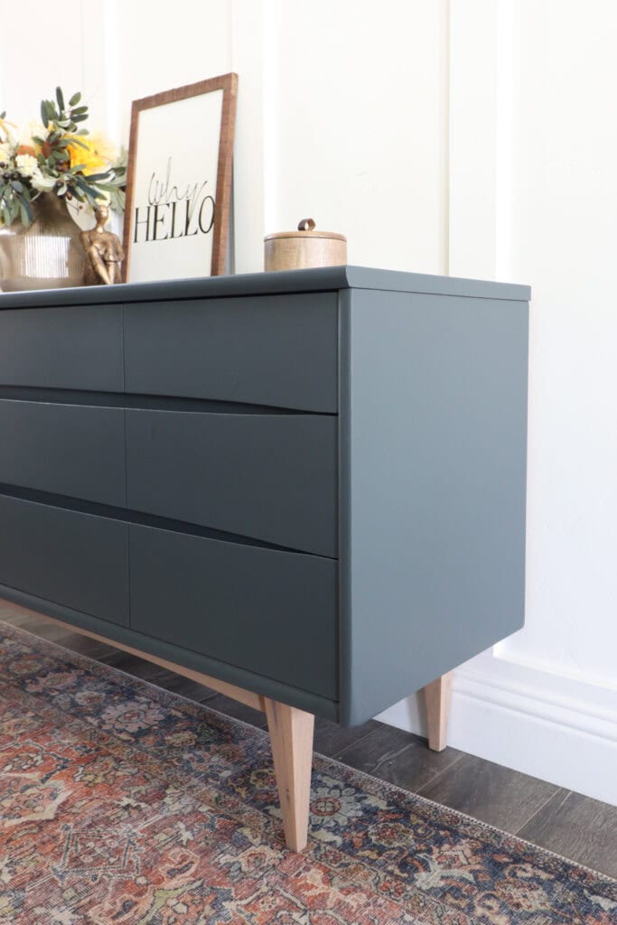 A modern blue dresser with wooden legs, topped with decor and a framed Hello sign on a patterned rug.